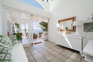 Quarto claro e ensolarado com vista para o mar, com um sofá branco, uma mesa de jantar e um espelho grande.