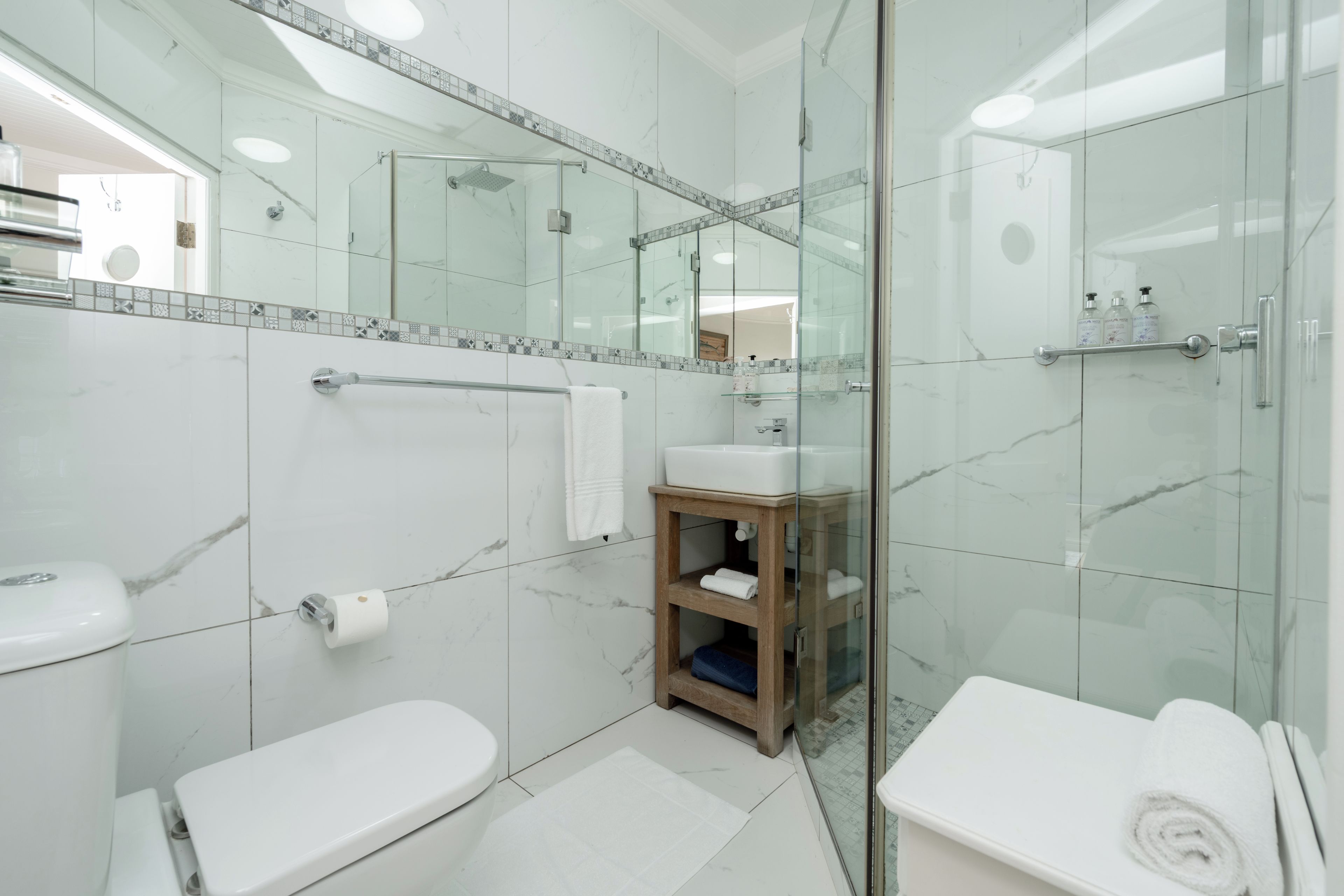 Un baño moderno con cabina de ducha de vidrio, inodoro blanco, lavamanos con tocador de madera y puerta de vidrio.