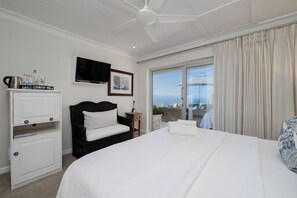 A hotel room with a bed, a television, a small table with a plant, and a view of the sea through the window. - Ocean Watch Guest House (Plettenberg Bay)