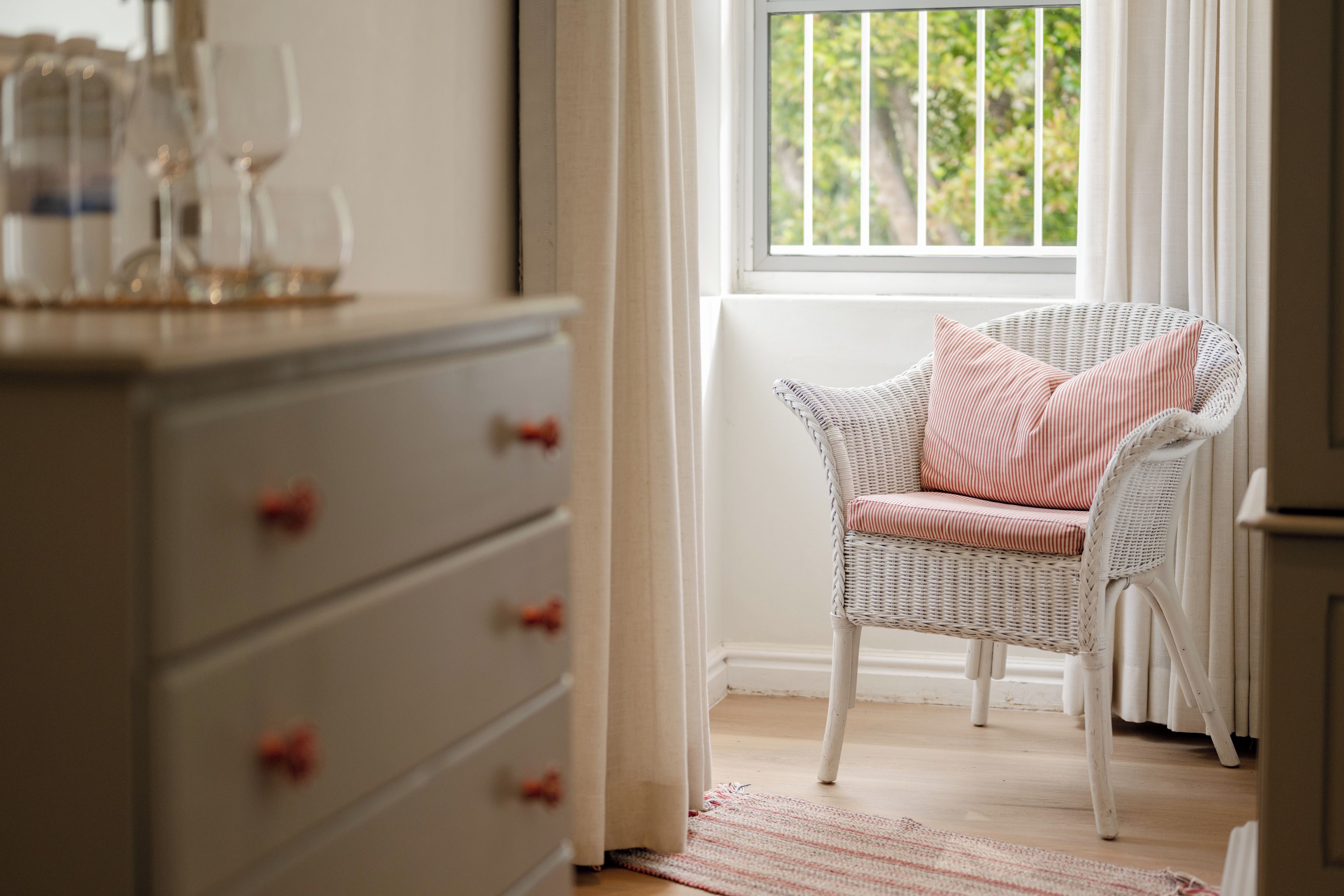 A room with a wicker chair, a white dresser with red knobs, and a window with a view of greenery.