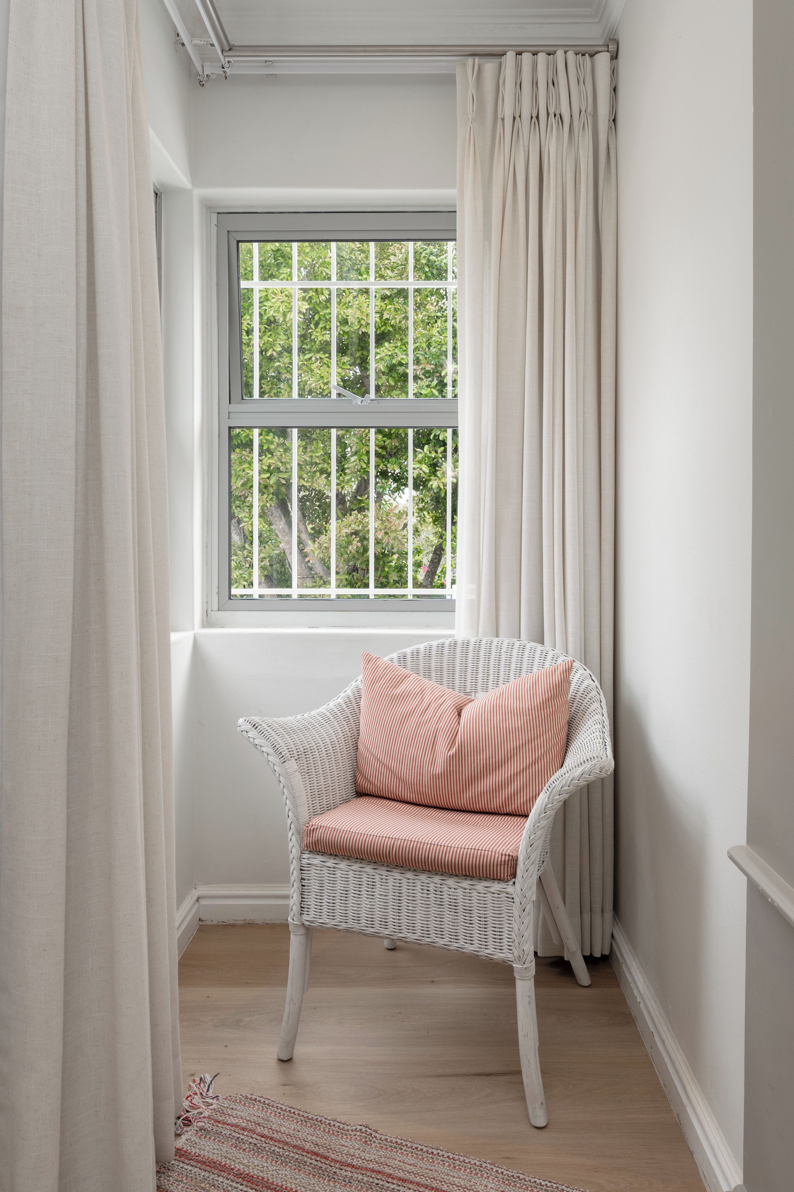 Habitación de esquina con una silla de mimbre blanca, una ventana con vista a follaje y una cortina blanca.