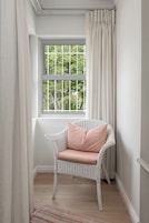 A corner room with a white wicker chair, a window with a view of greenery, and a white curtain.