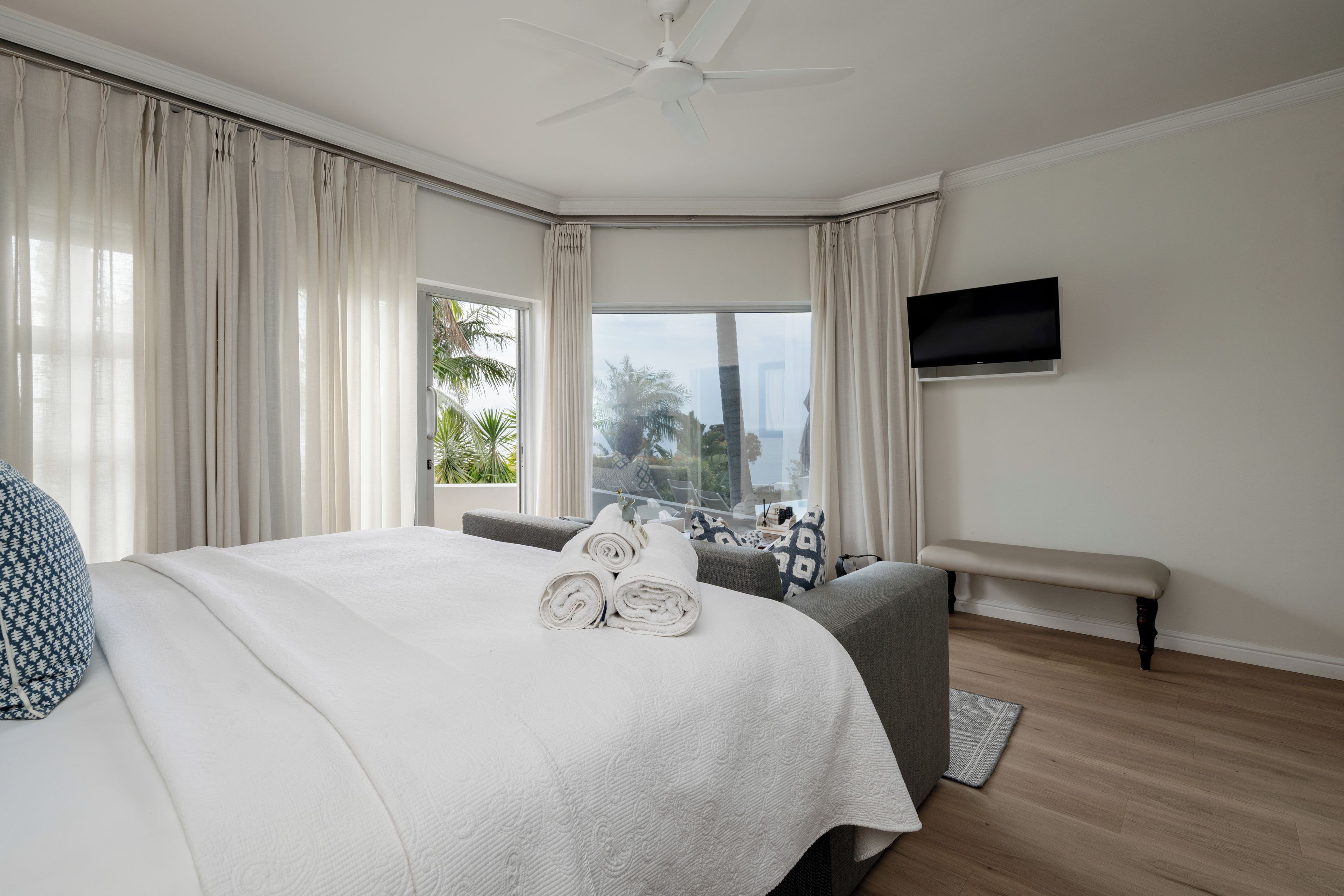 Habitación de hotel con cama, un banco, televisión de pantalla plana y vistas a palmeras y edificios.