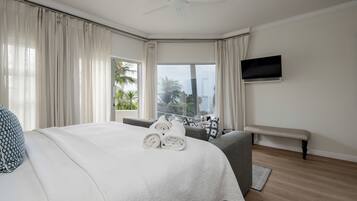 Habitación de hotel con cama, un banco, televisión de pantalla plana y vistas a palmeras y edificios.