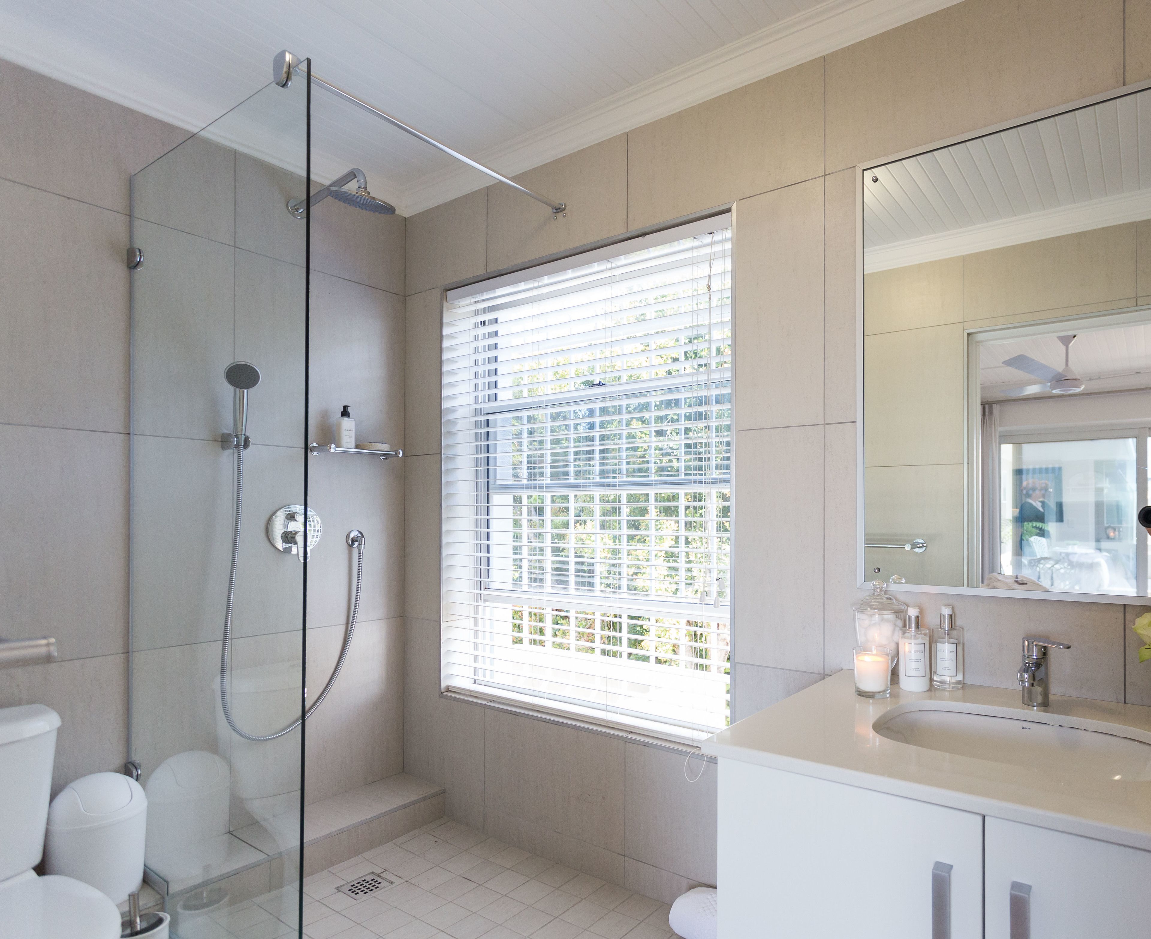 Un baño moderno con ducha de vidrio, un ventanal con persianas, un lavamanos blanco y un espejo.