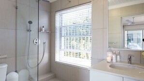 A modern bathroom with a glass-enclosed shower, a large window with blinds, a white sink, and a mirror. - Ocean Watch Guest House (Plettenberg Bay)