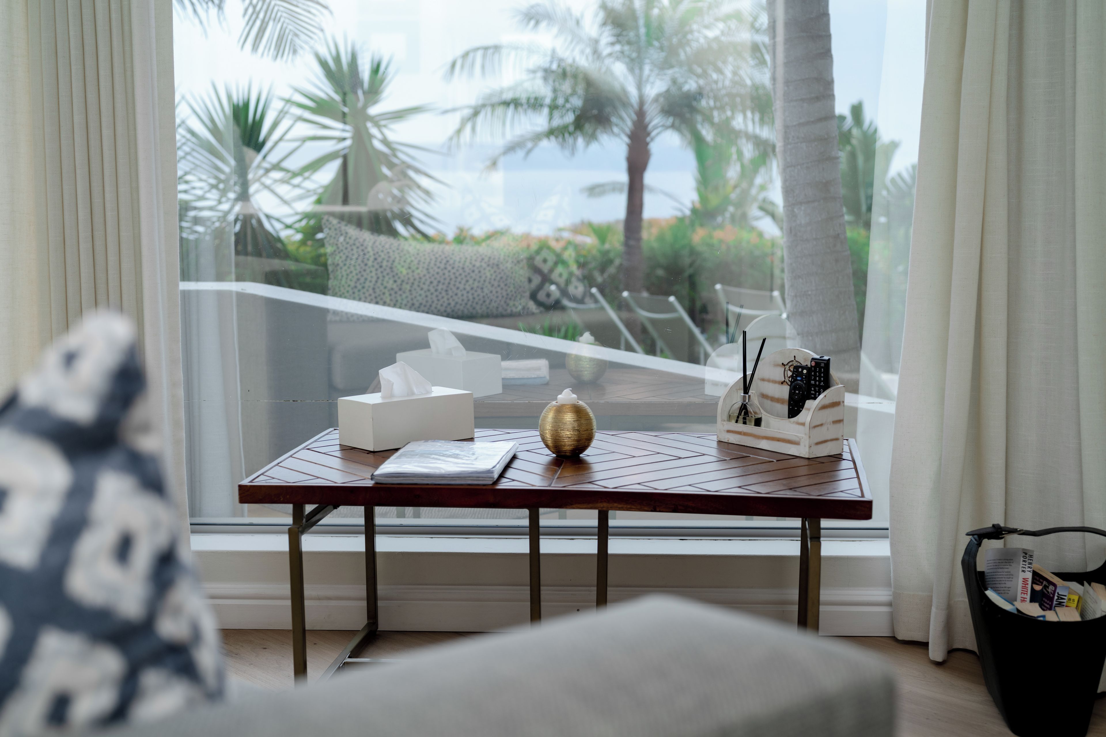 A room with a view of palm trees and the ocean through a large window. There is a table with a tissue box, a decorative bowl, and a remote control holder.