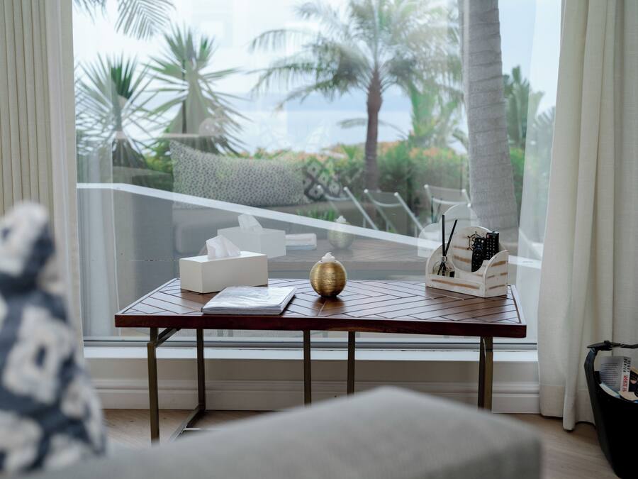 A room with a view of palm trees and the ocean through a large window. There is a table with a tissue box, a decorative bowl, and a remote control holder.