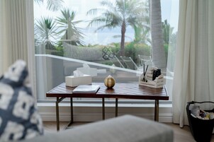 Una habitación con vistas a palmeras y al océano a través de un ventanal grande. Hay una mesa con una caja de pañuelos, un cuenco decorativo y un soporte para control remoto.