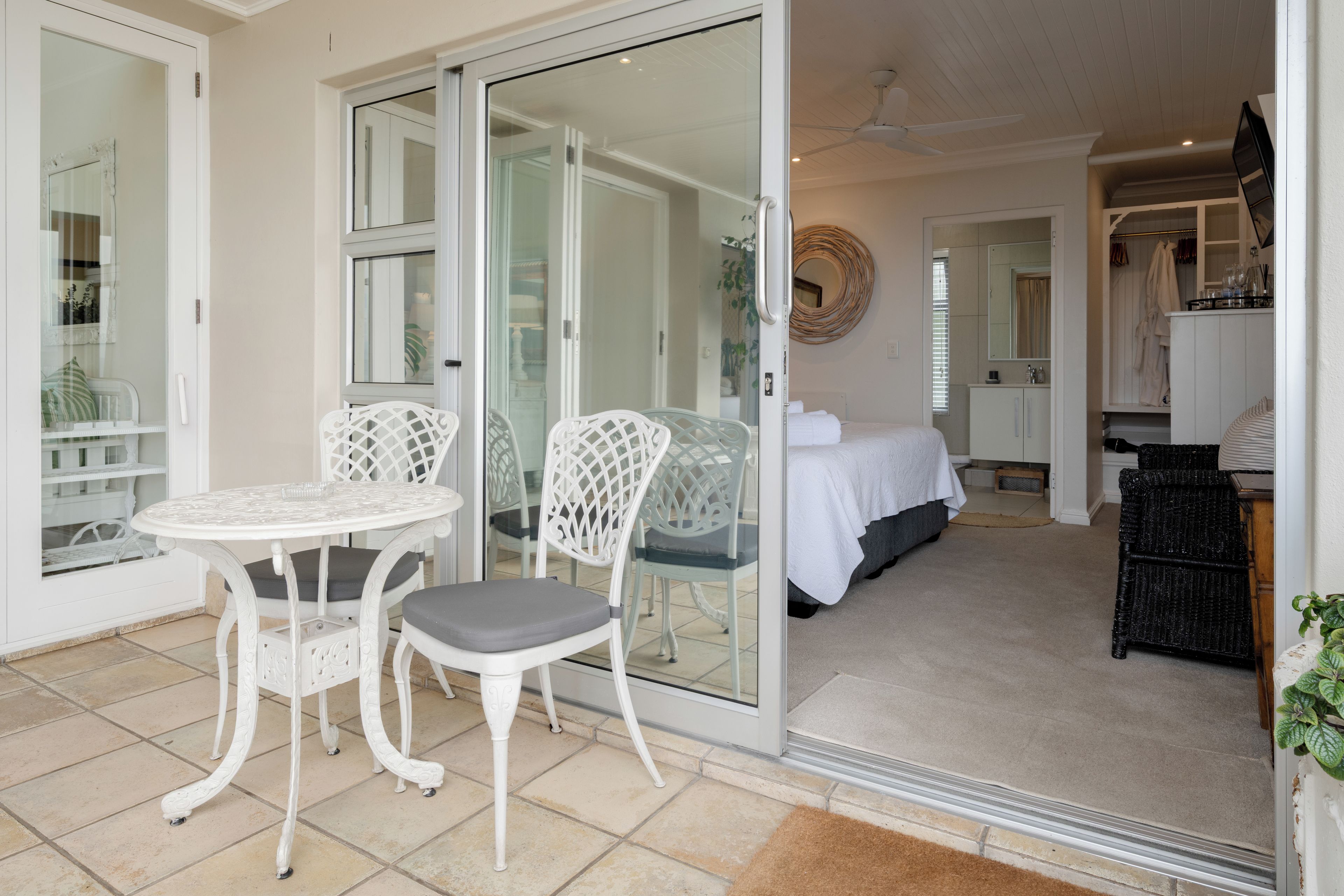 Una habitación con puerta de cristal que da a un balcón, una cama con colcha blanca, una mesa de comedor con sillas y una pequeña cocina.