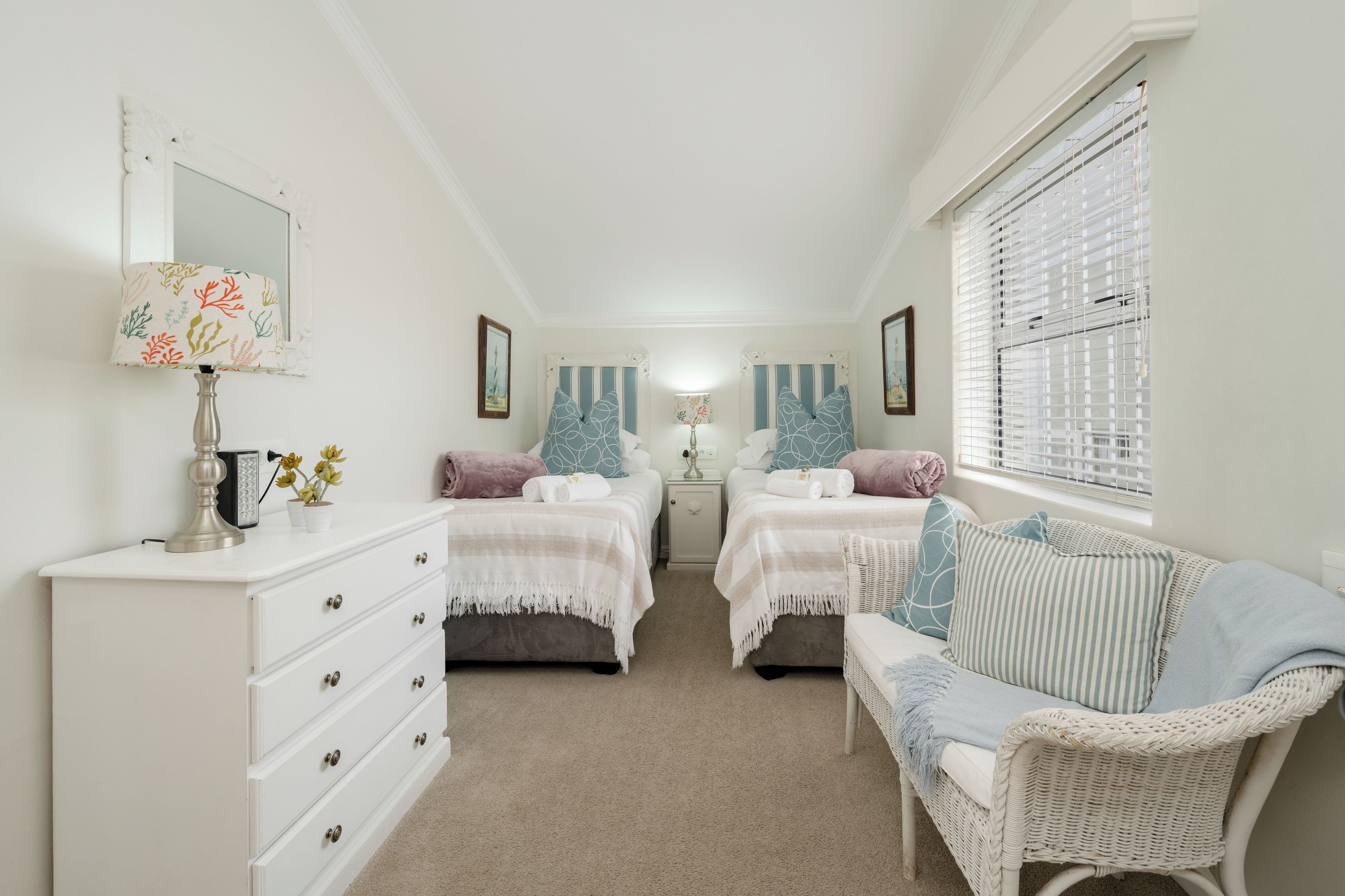 A narrow hallway with two beds, a white dresser, and a wicker chair.
