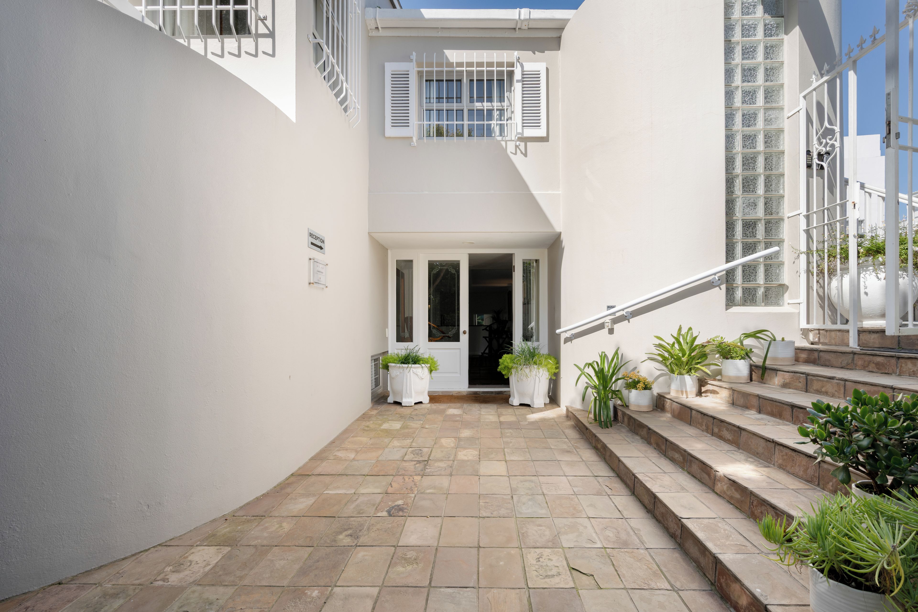 Un estrecho patio con una escalera que conduce a una puerta, rodeado de paredes blancas y plantas en macetas.