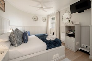 A bedroom with a bed, bedside tables, a wardrobe, a ceiling fan, a window with curtains, and a mounted television. - Ocean Watch Guest House (Plettenberg Bay)