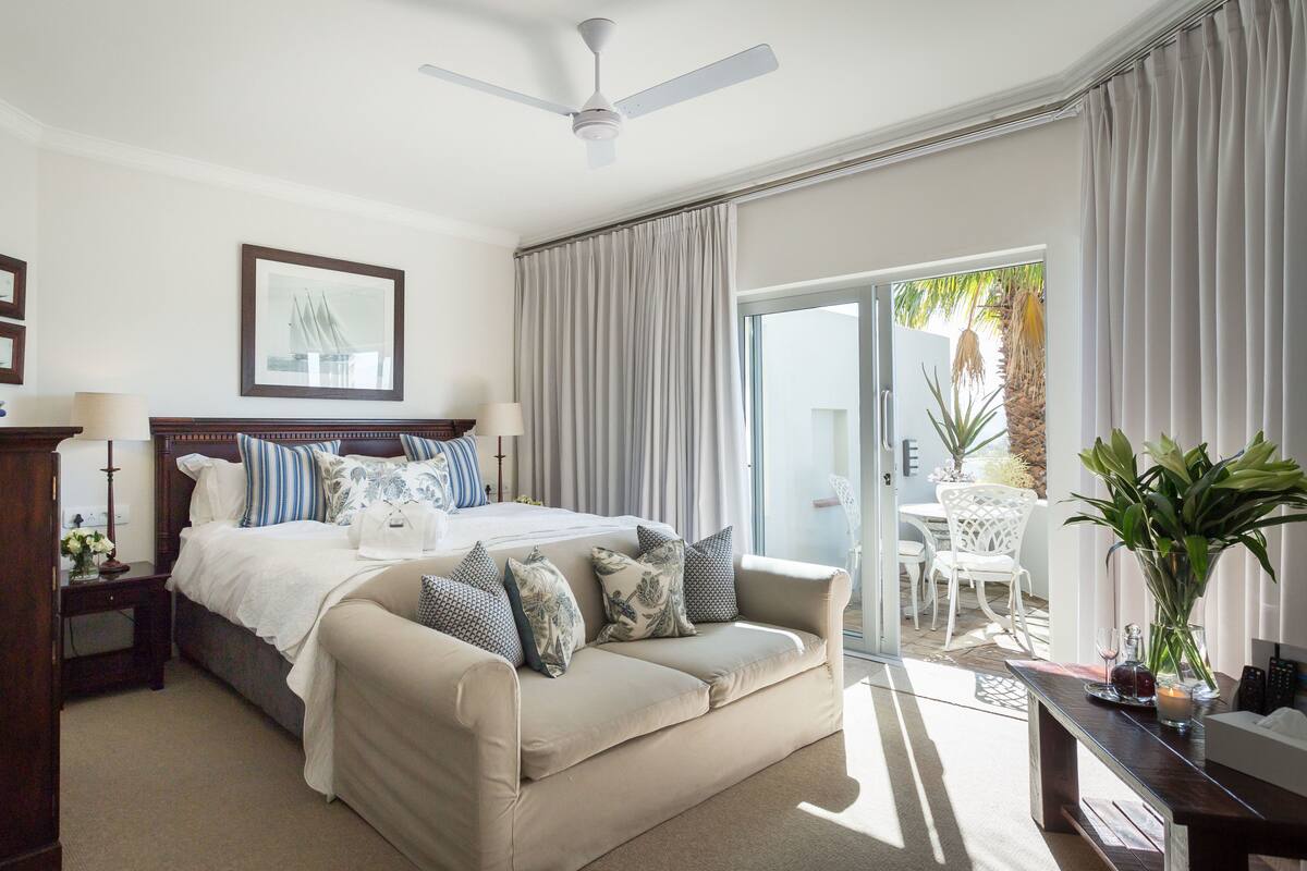 A bedroom with a bed, a sofa, a ceiling fan, a large window with curtains, and a view of the outdoors.