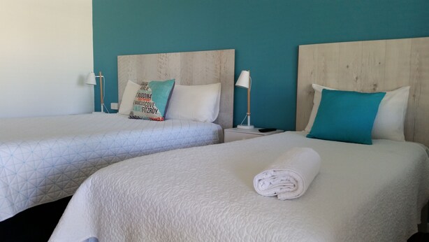 Deux lits jumeaux avec une literie blanche et des oreillers turquoise, une tête de lit en bois, une table de chevet avec une lampe et une serviette roulée posée sur le lit.