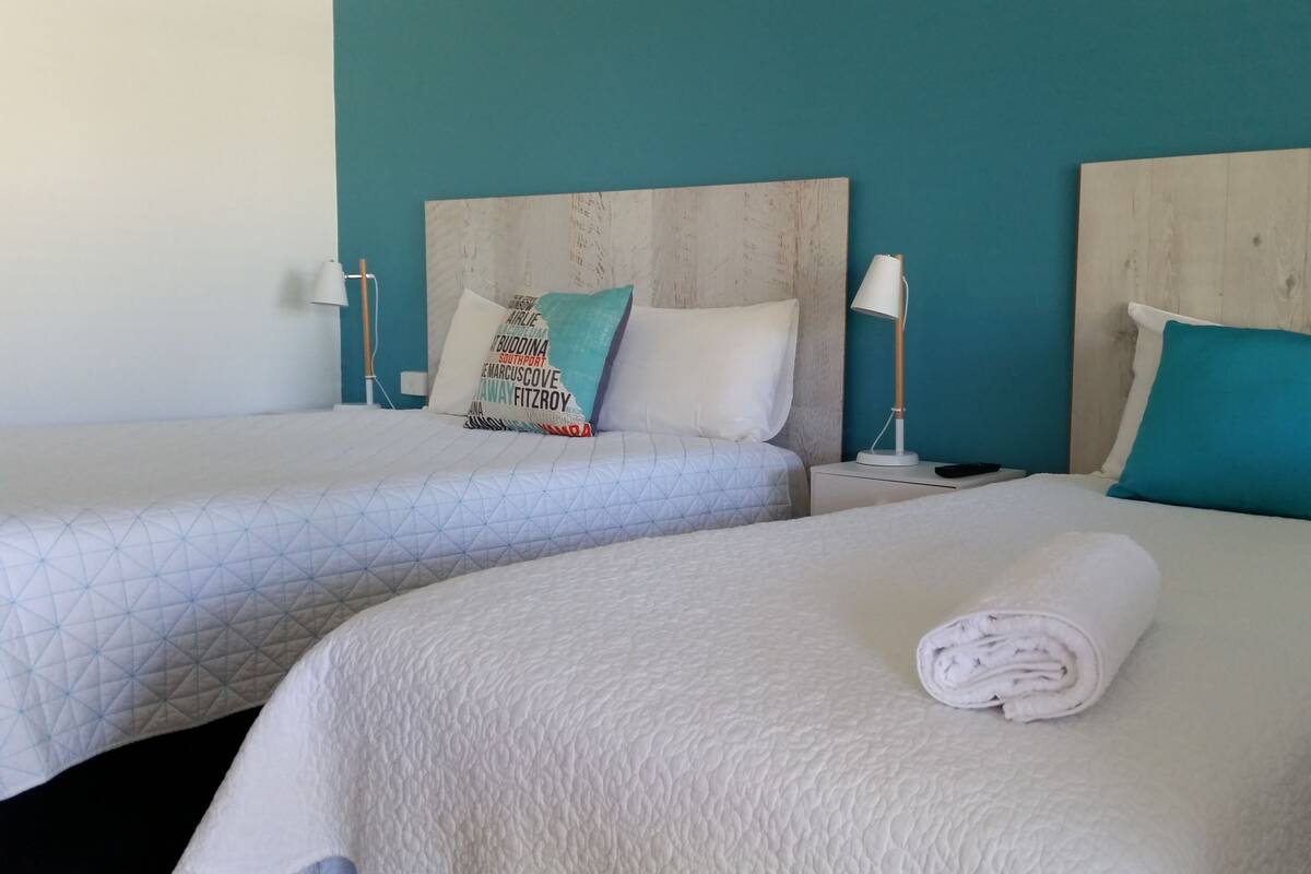 Two twin beds with white bedding and teal pillows, a wooden headboard, a bedside table with a lamp, and a rolled towel on the bed.