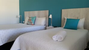 Zwei Einzelbetten mit weißer Bettwäsche und türkisfarbenen Kissen, ein Holz-Kopfteil, ein Nachttisch mit Lampe und ein zusammengerolltes Handtuch auf dem Bett.