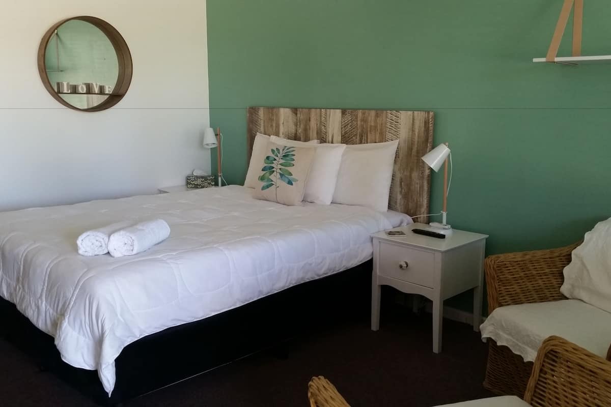 A neatly made bed with white linens and rolled towels, a bedside table with a lamp, and a wicker chair in a room with green walls and a mirror on the wall.