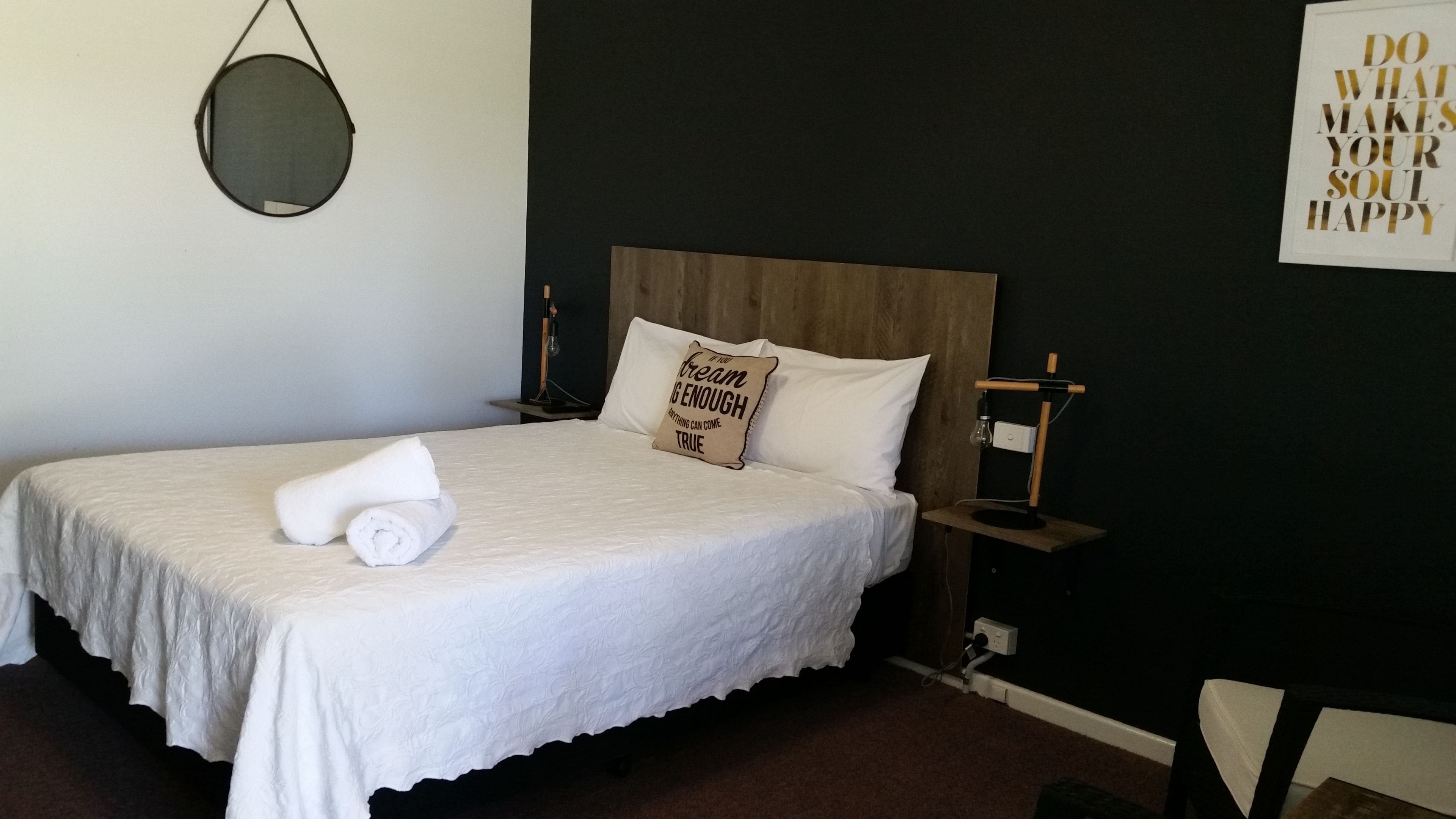 A neatly made bed with white linens, a pillow with a motivational quote, a bedside table with a lamp, and a wall with a motivational poster.