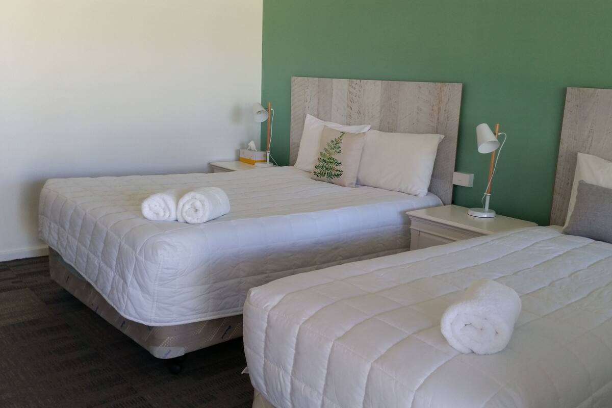 A hotel room with two beds, each with a white duvet and rolled towels. There are bedside tables with lamps and a small plant on one bed.