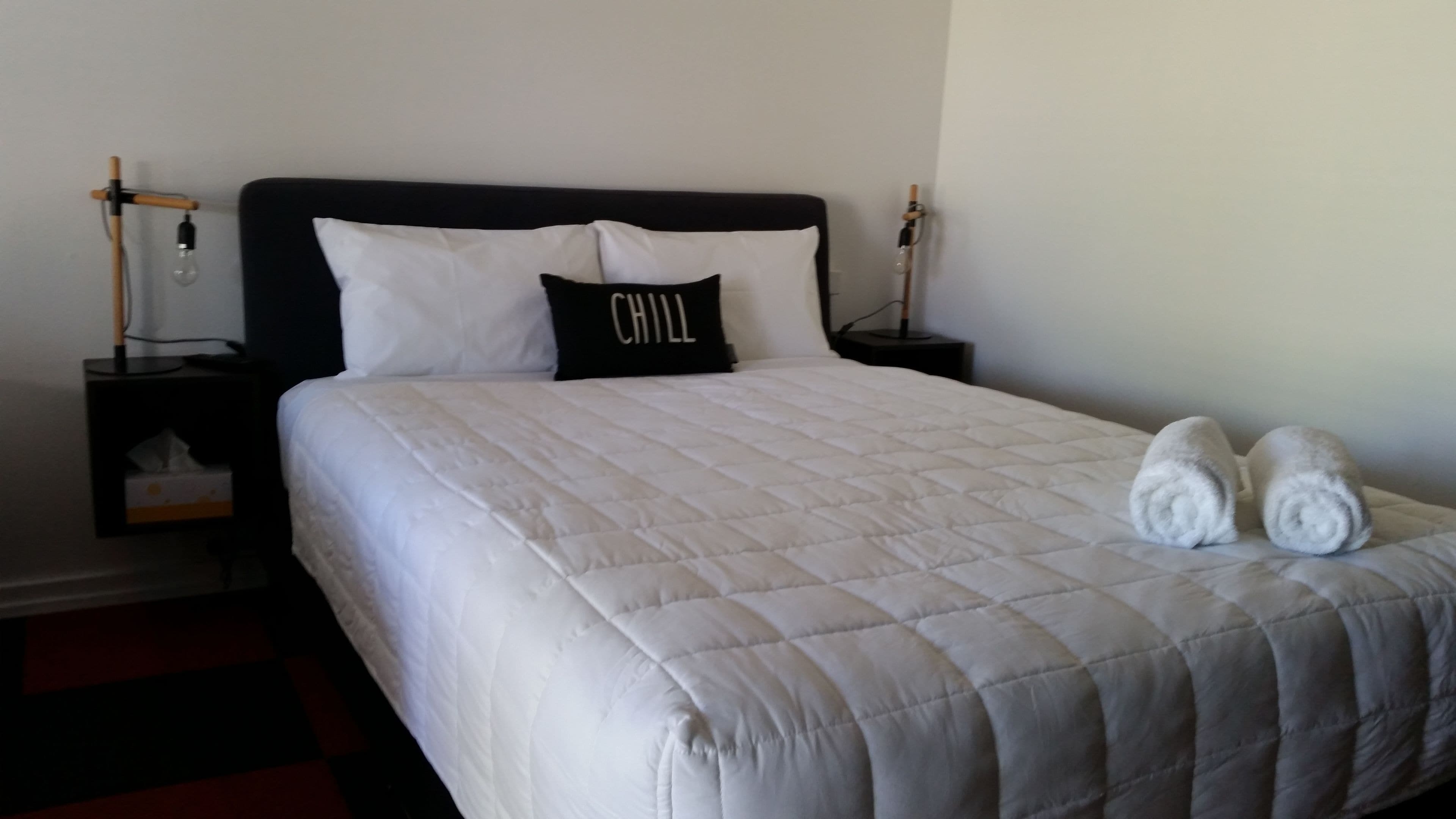 A neatly made bed with a white quilted bedspread, two pillows, and two rolled towels on the foot of the bed.