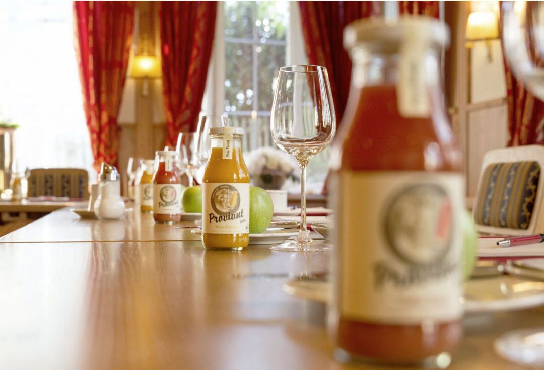 Ein Tisch mit Proviant-Weinflaschen, Gläsern, einem grünen Apfel und einer Wasserflasche.