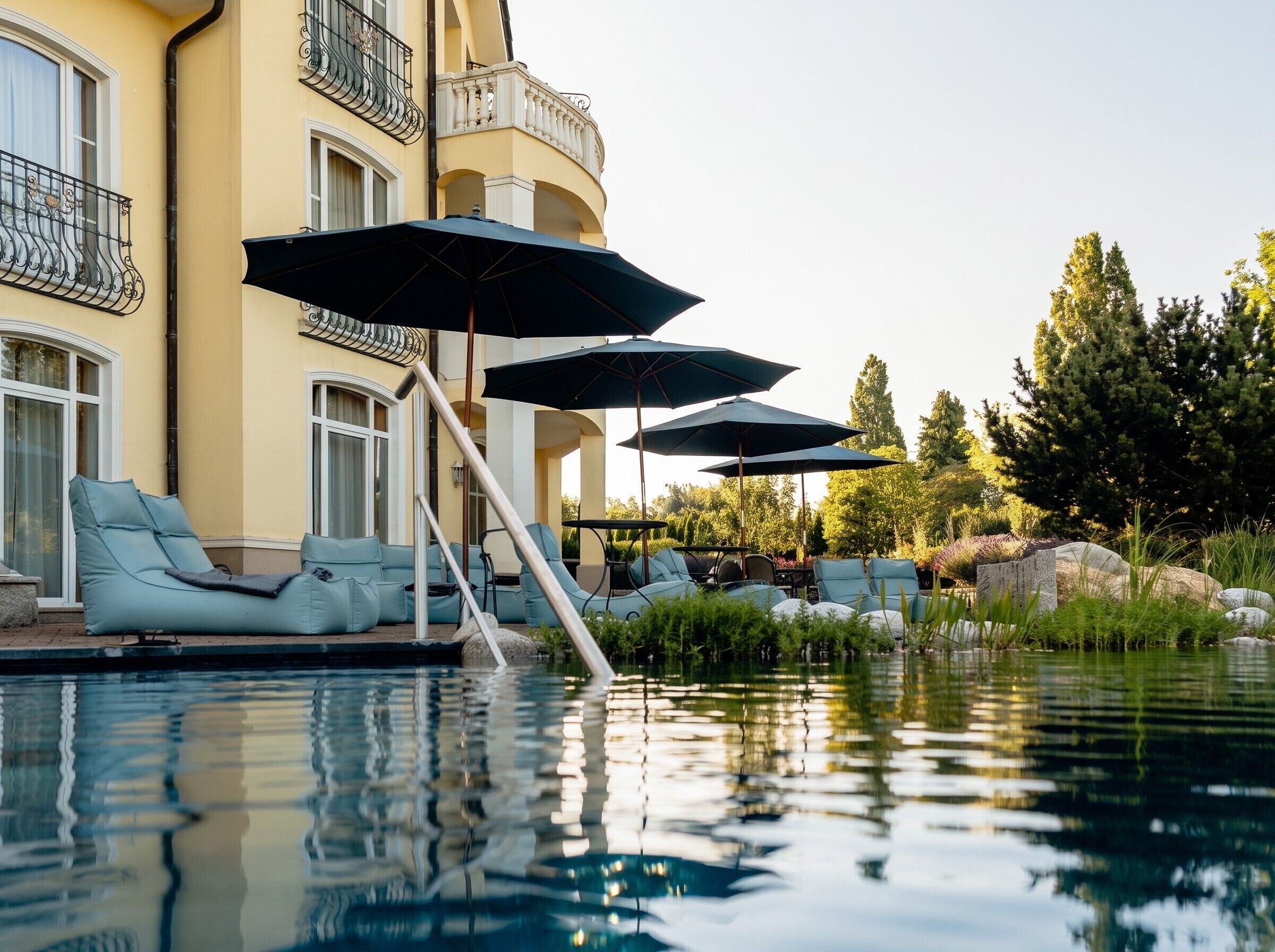 Zona piscina con lettini e ombrelloni, un edificio con balconi e un riflesso nell'acqua.