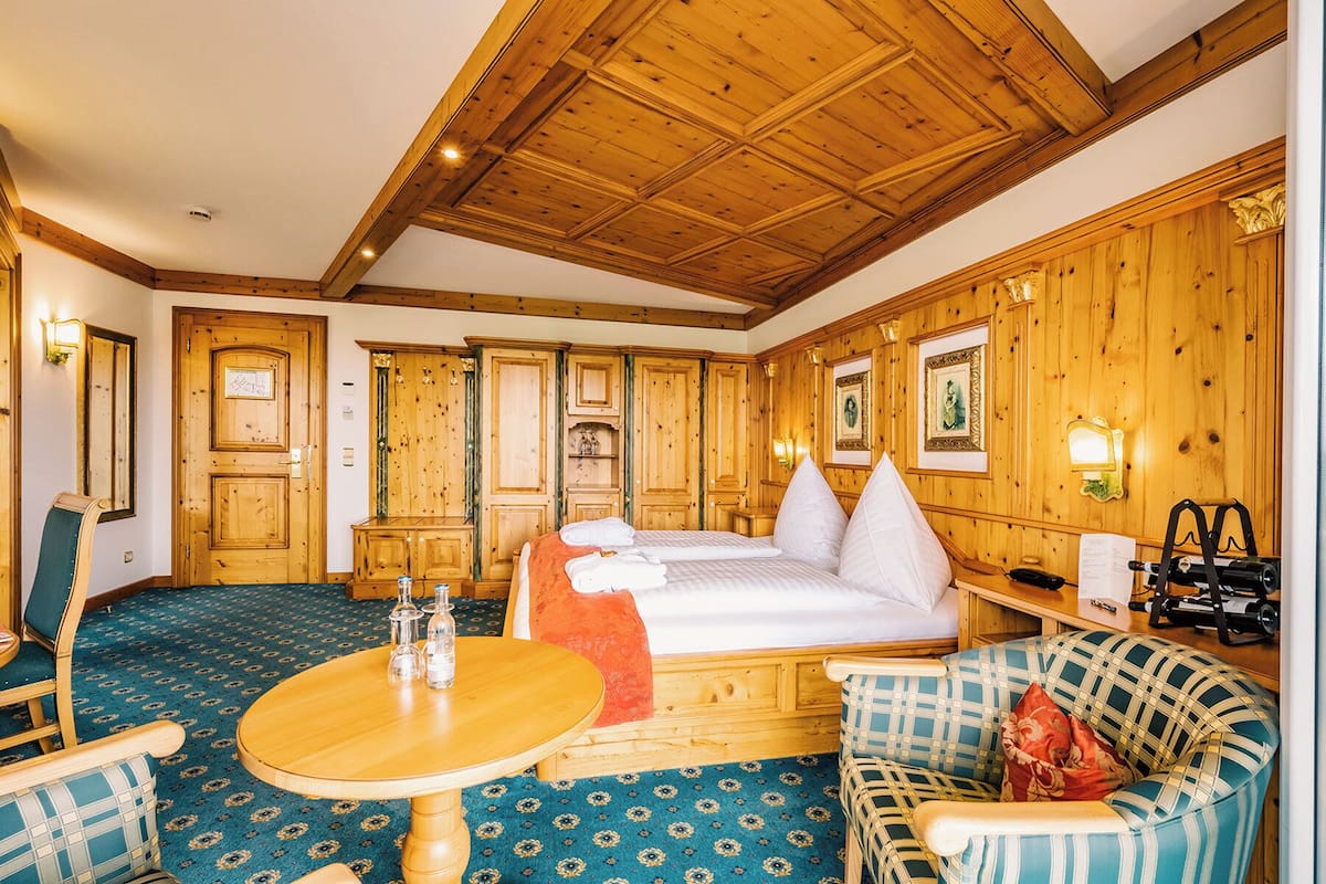 Een kamer met een houten plafond, een bed met wit beddengoed, een ronde houten tafel met zout- en peperstel, een geruit sofa en een kamerbreed tapijt met patroon.