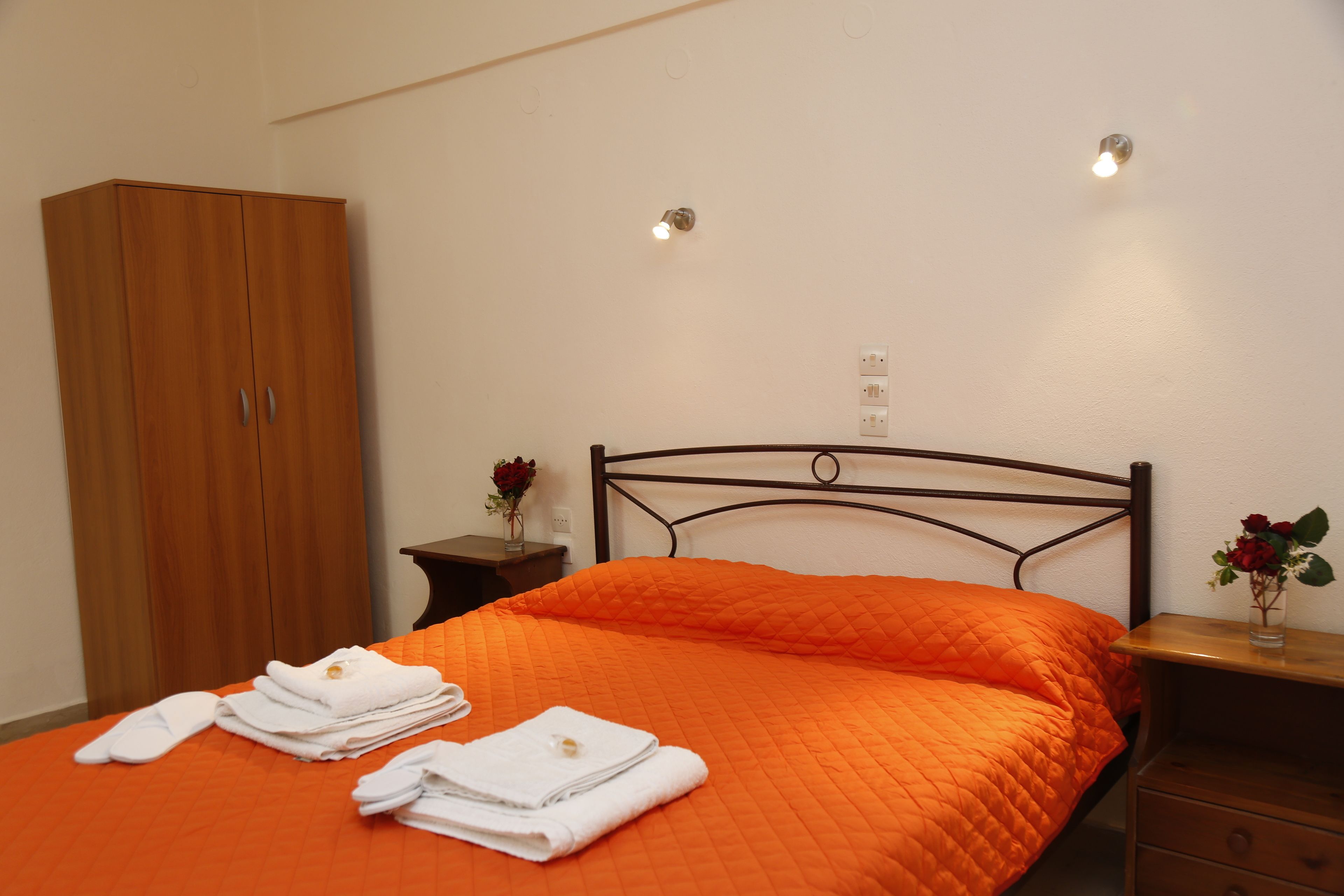 Hotellihuone, jossa on sänky, puinen vaatekaappi, yöpöytä maljakkoineen ja seinälle asennettu valaisin.