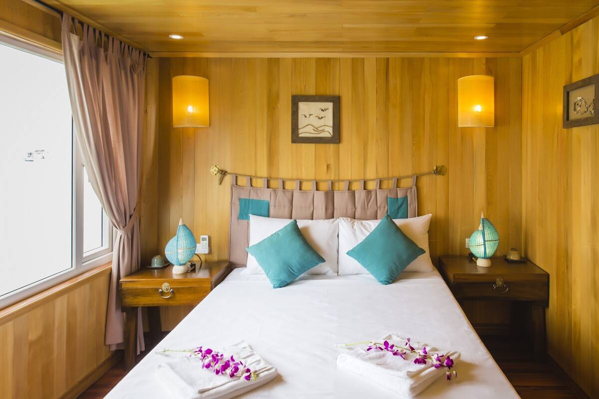 Une chambre d’hôtel avec un mur lambrissé en bois, un lit avec une literie blanche et des coussins turquoise, deux lampes de chevet, une table de chevet avec un globe terrestre et une lampe, et une fenêtre avec des rideaux.
