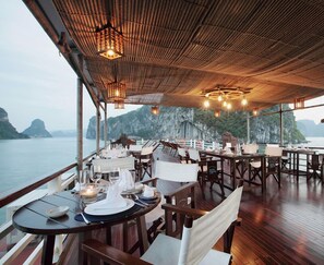 Un restaurant dont les tables sont dressées pour le service, offrant une vue sur un plan d’eau et des falaises.