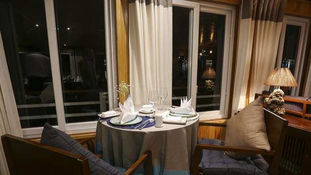 A dining area with a round table set for two, a vase with flowers, and a lamp on a side table. - Secret Halong Cruise (Bai Chay)