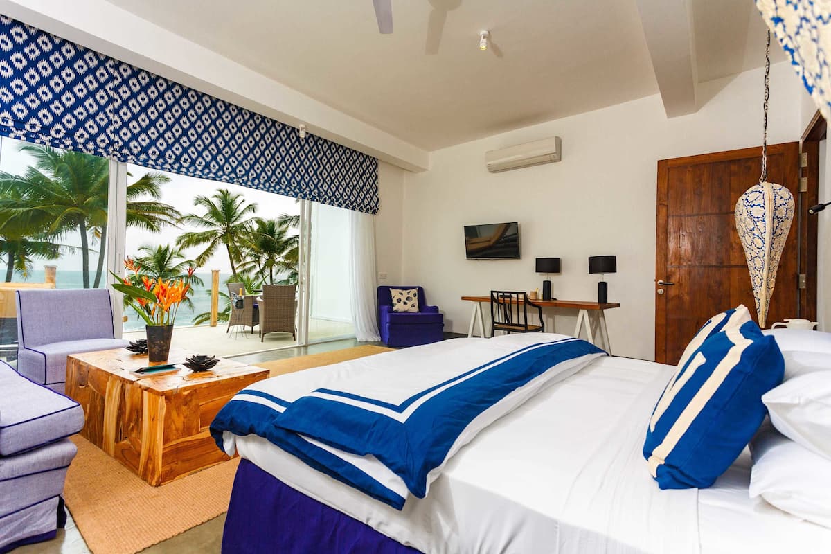 A hotel room with a bed, a wooden coffee table, a purple armchair, a desk, and a view of palm trees and the sea.