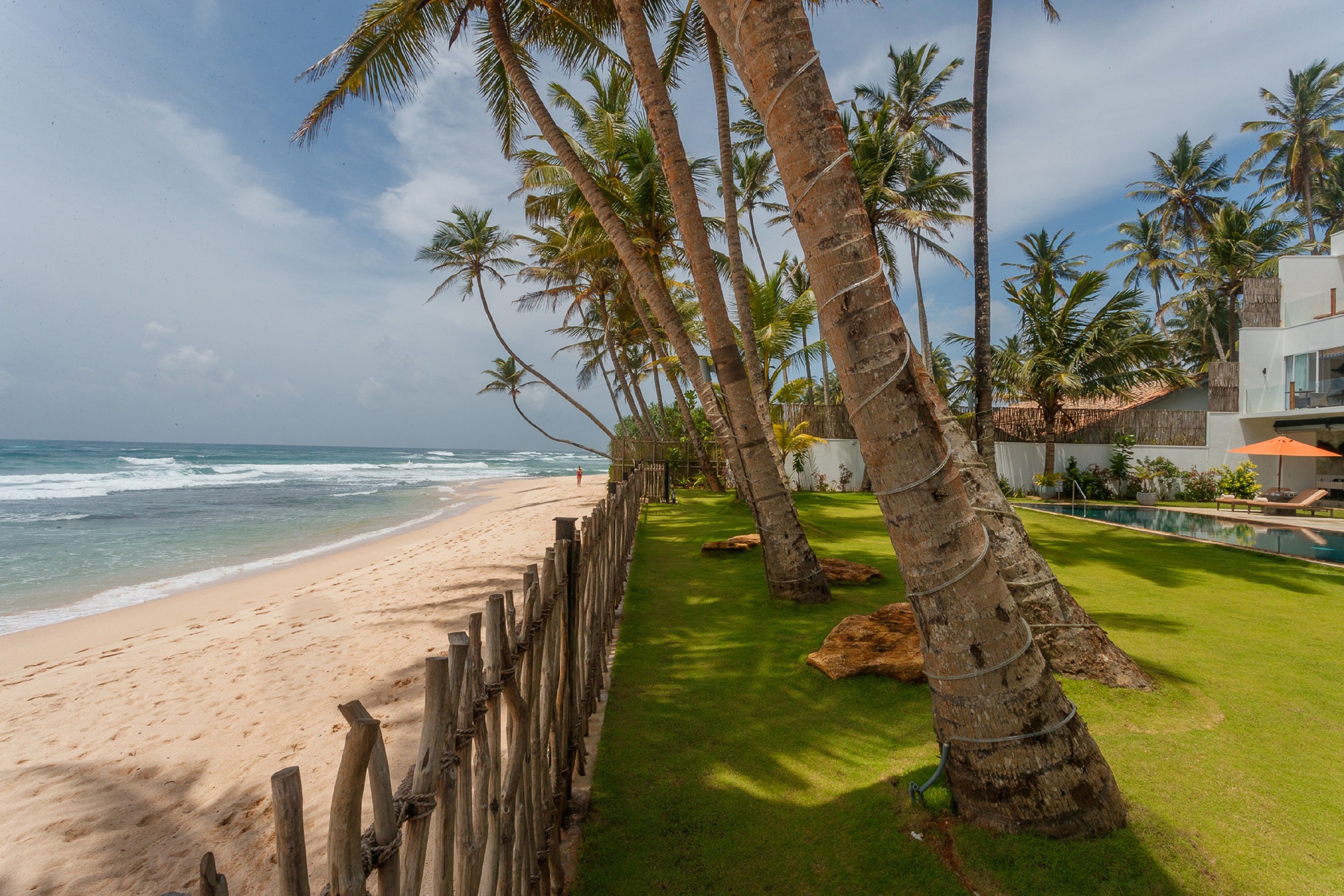 A beach with a wooden fence, palm trees, and a modern building with a pool.