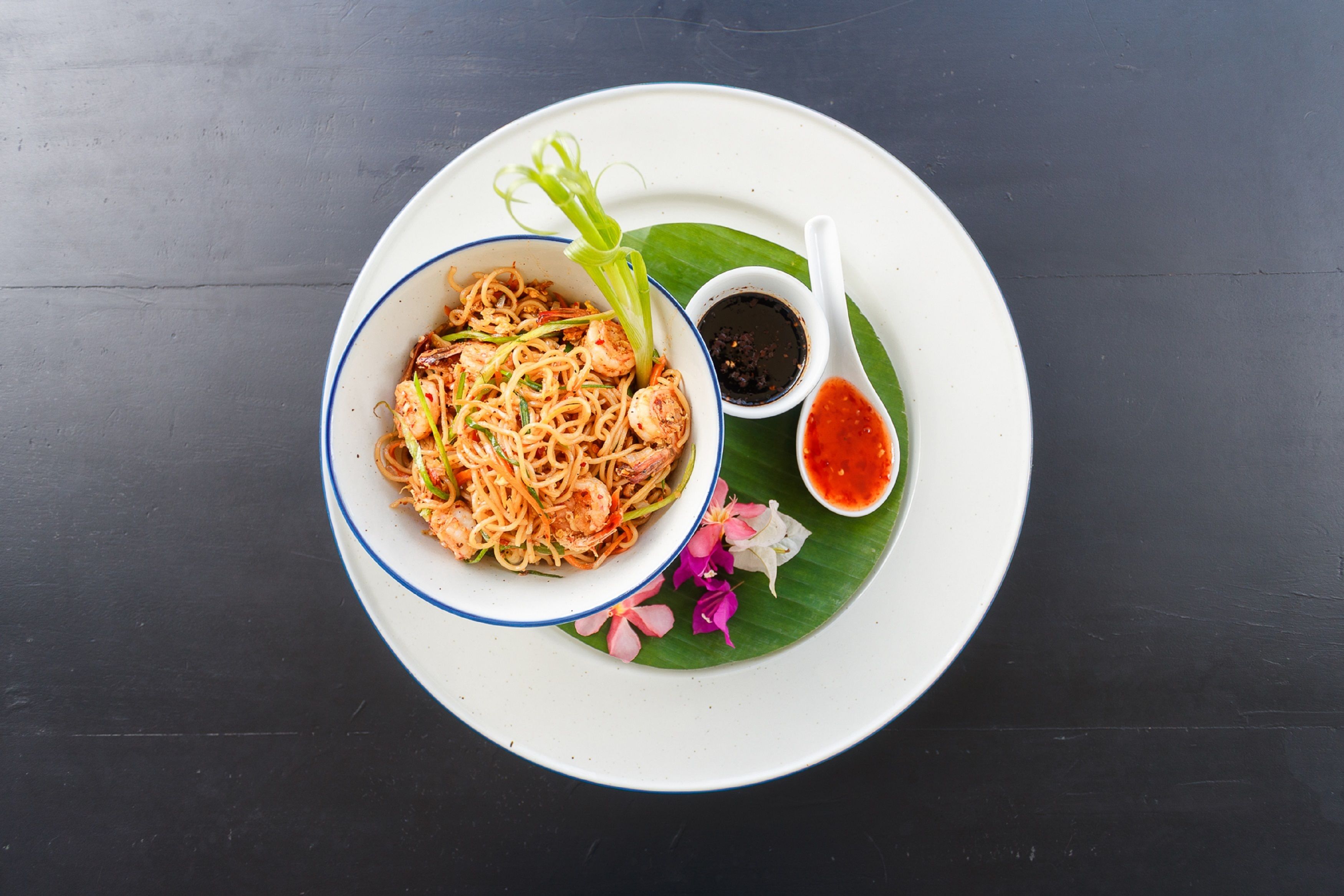 A bowl of noodles with shrimp and vegetables, served on a white plate with a banana leaf and a spoon of sauce.