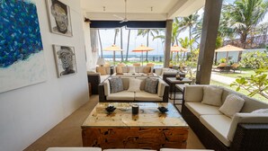 Una sala de estar moderna con un ventanal grande que da a una playa, amueblada con un sofá, una mesa de centro y cojines decorativos.