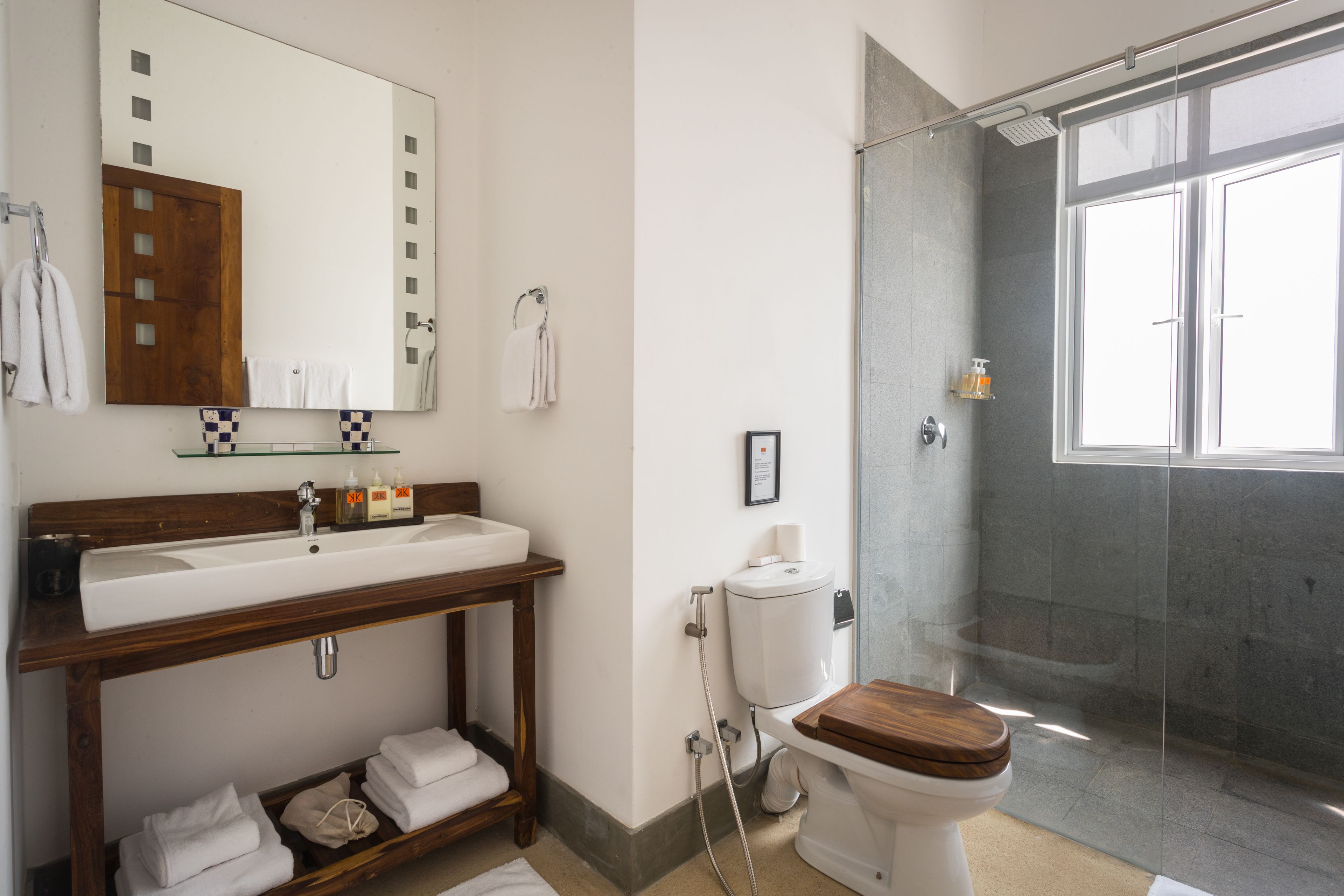 A modern bathroom with a wooden vanity, a large mirror, a glass shower enclosure, and a toilet with a wooden seat.