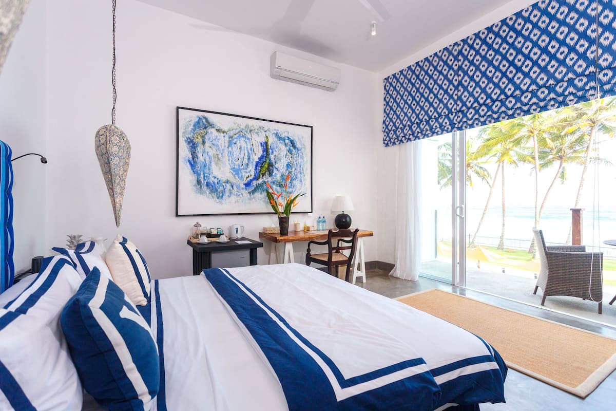 A hotel room with a bed, desk, chair, and a large window with a view of palm trees and the ocean.