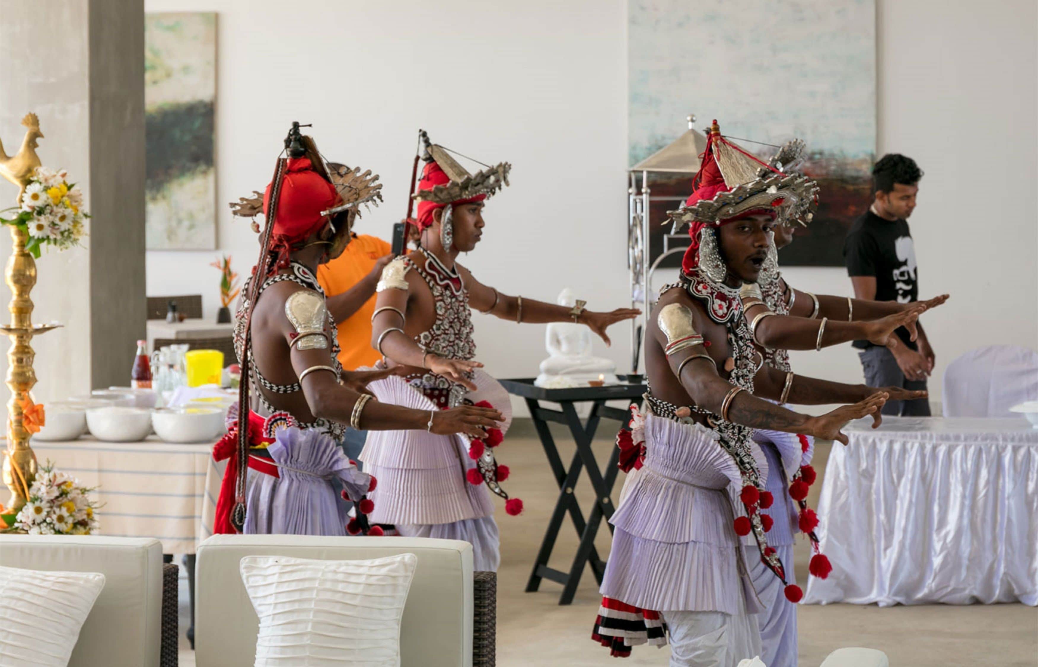 Un grupo de personas vestidas con atuendo tradicional realizan una danza en una sala con sillas blancas y una mesa decorada con arreglos florales.