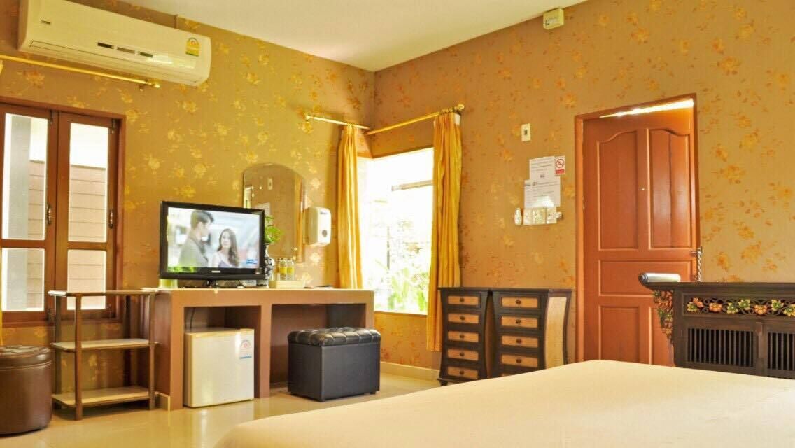 A hotel room with a bed, a television, a desk with a computer, a small refrigerator, a trash bin, and a window with curtains.