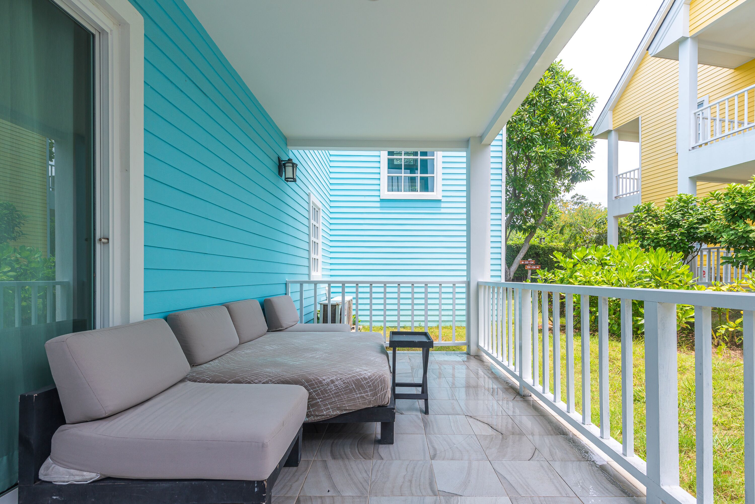 A covered porch with a beige sofa and a small table, a white railing, and a blue building in the background.