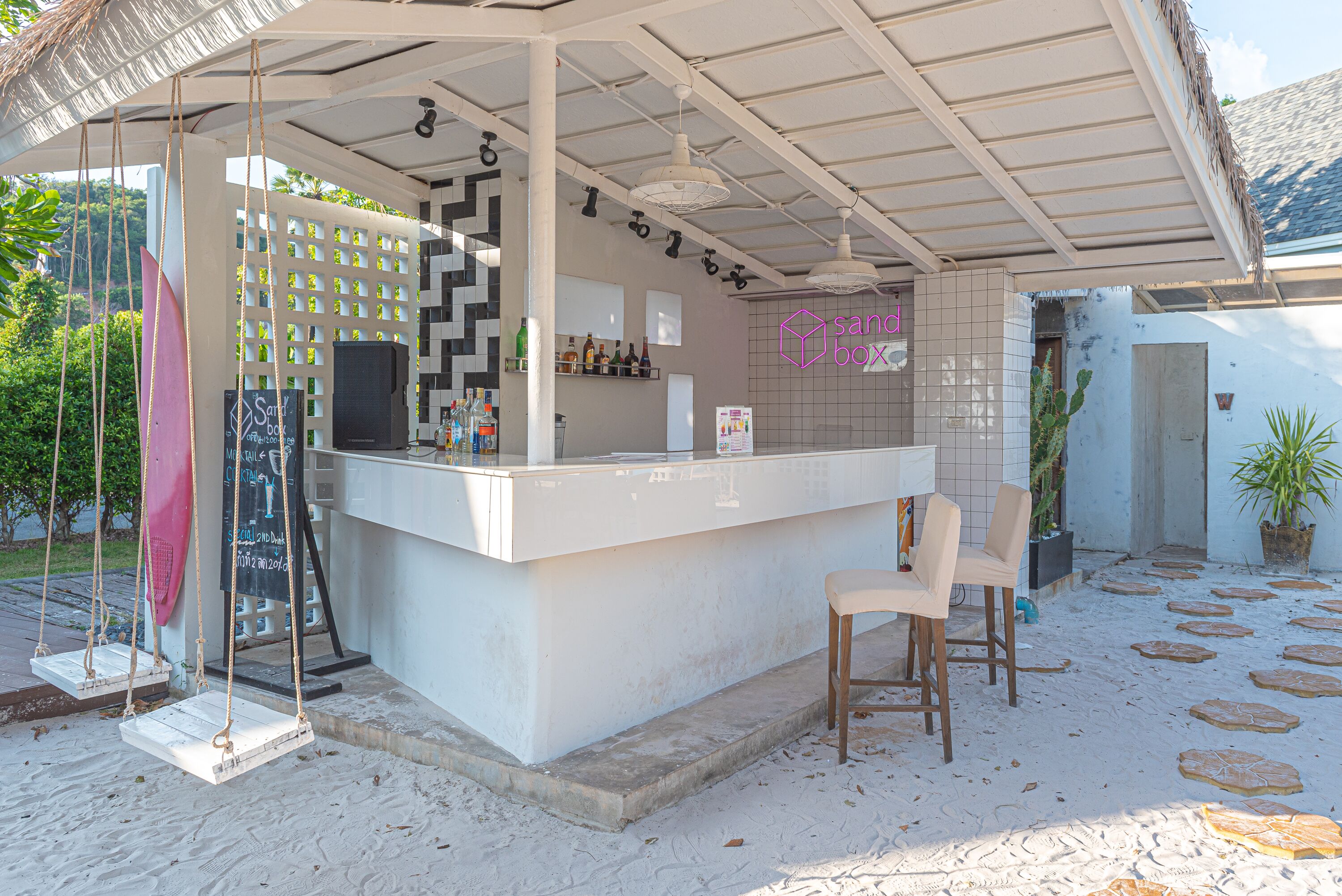 A beachside bar with a white counter, two bar stools, and a swing chair.