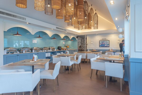 Un restaurant moderne avec des chaises et des tables blanches, un comptoir où sont exposés des produits alimentaires, et des luminaires suspendus en osier.