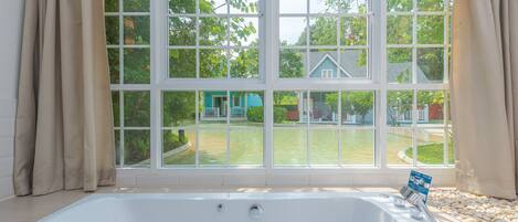 A large, white bathtub with multiple faucets and a control panel, positioned in front of a large window with a view of a house and lawn.