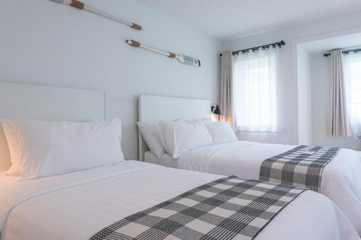 Twee bedden met witte lakens, een hoofdbord en een geruit plaid.