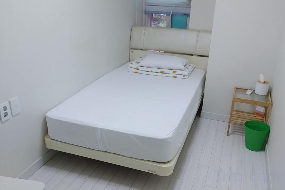 A single bed with a white mattress and a pillow, a wooden bedside table with a lamp and a green trash bin, and a window with curtains.
