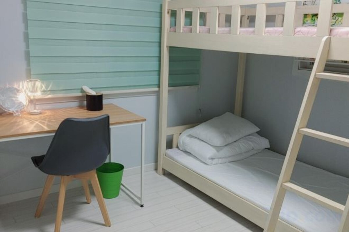 A bunk bed with a desk and chair, a green trash bin, and a window with blinds.