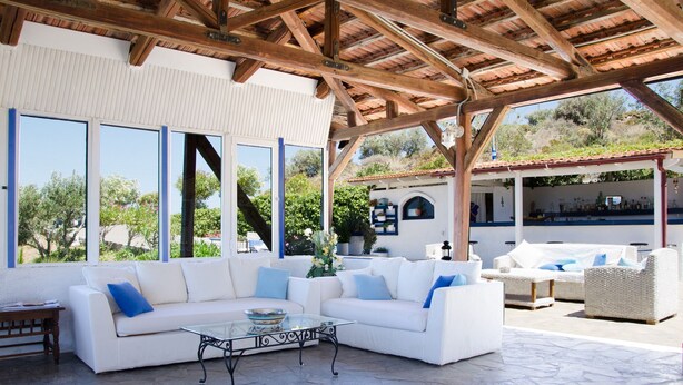 A covered outdoor seating area with a white sofa and a glass coffee table, stone flooring, and a view of a bar area in the background. - Bellair Village (Malevizi)