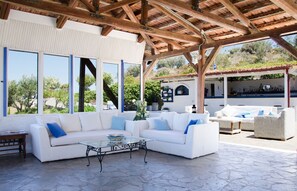 A covered outdoor seating area with a white sofa and a glass coffee table, stone flooring, and a view of a bar area in the background. - Bellair Village (Malevizi)