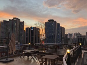 Terraza en la azotea con mesas y sillas, con vistas a un paisaje urbano con edificios altos al atardecer.